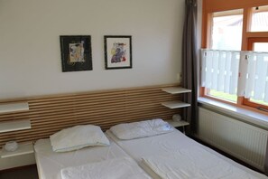 A hotel room with a wooden headboard, two beds, a window with blinds, and two framed pictures on the wall. - Bungalow with 2 bedrooms (Julianadorp aan zee)