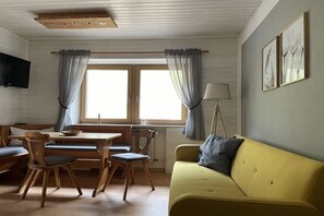 Ein Zimmer mit einem Holztisch, Stühlen, einem gelben Sofa, einer Stehlampe und einem an der Wand montierten Fernseher.