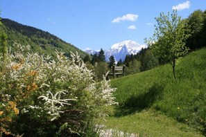 Eine Berglandschaft mit blauem Himmel, einem Weg zu einer Holzhütte und schneebedeckten Bergen im Hintergrund.