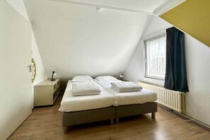 Ein Einzelbett mit weißer Bettwäsche in einem Zimmer mit schrägen Wänden und einem fensterbelaubten Fenster.