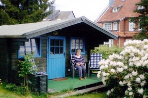 Een klein houten gebouw met een blauwe deur en ramen, een veranda met twee stoelen, en een tuin met witte bloemen.