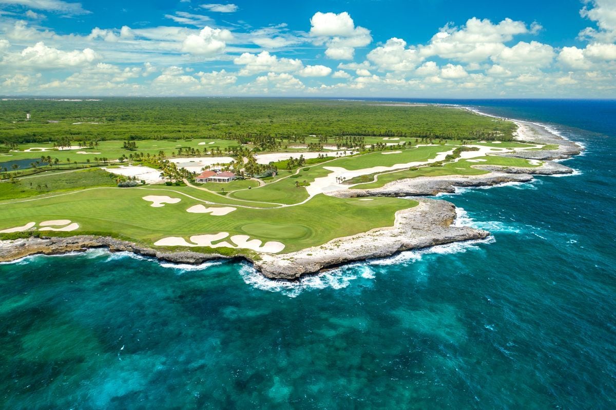 An aerial view of a coastal golf course with a clubhouse, sand bunkers, and a clear view of the ocean.
