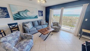 A living room with a sofa, armchair, and coffee table, a large painting of a wave, and a balcony with a view of the beach. - Pelican Isle 106 1 Bedroom Condo (Fort Walton Beach)