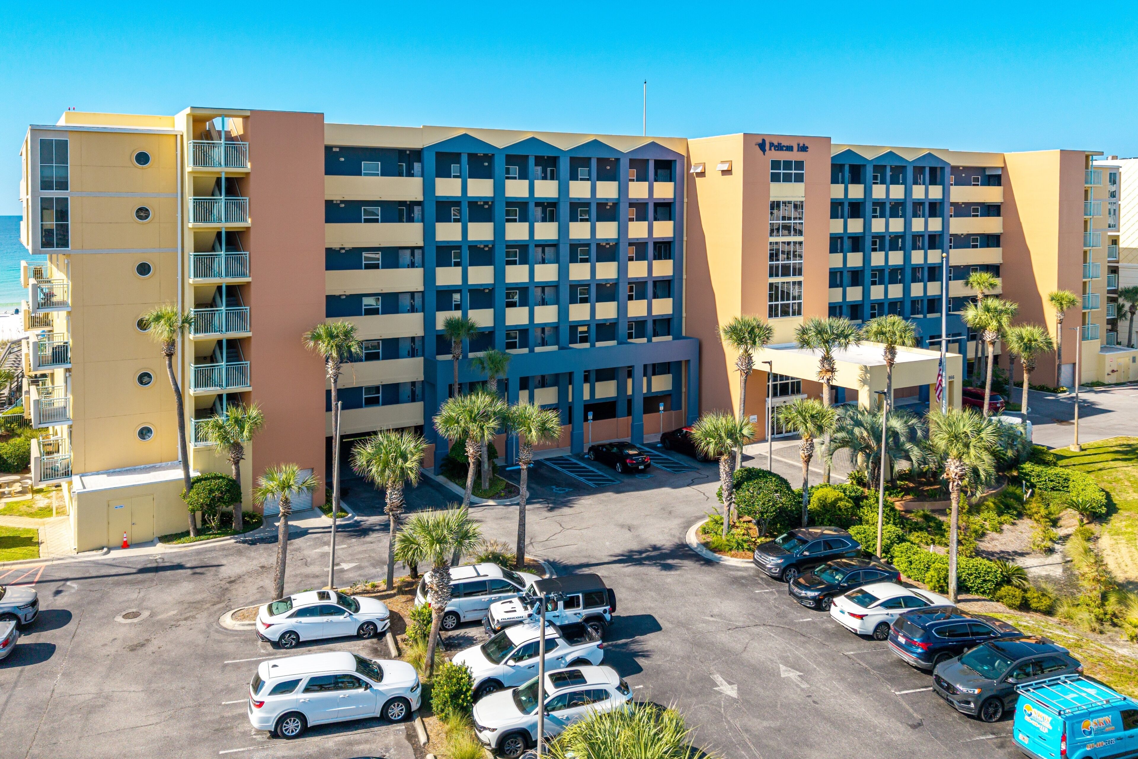 A multi-story hotel with a parking lot filled with cars, surrounded by palm trees and a clear blue sky.