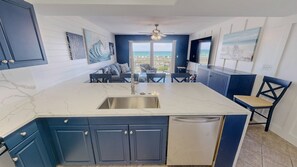 A modern kitchen with a large island, dark blue cabinets, and a view of the ocean through the windows. - Pelican Isle 106 1 Bedroom Condo (Fort Walton Beach)