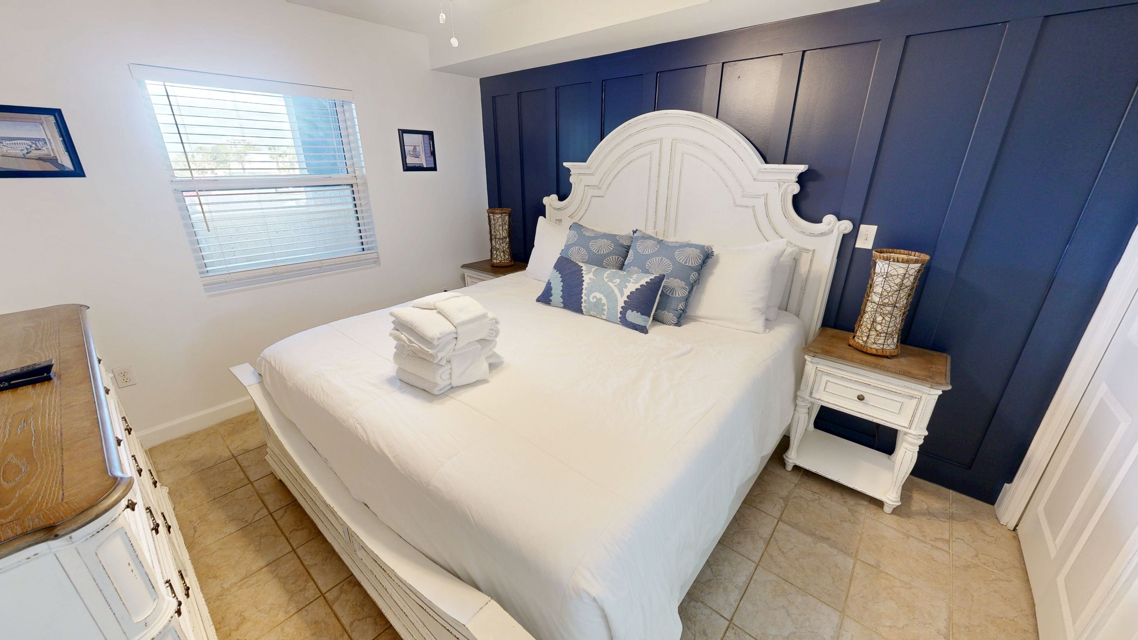 A neatly made bed with white linens and a stack of folded towels, a bedside table with a decorative vase, and a window with blinds.
