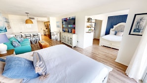 Quarto moderno com uma cama grande, área de estar com sofá e poltronas, cozinha ao fundo e TV fixada na parede.