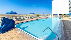 Área da piscina com espreguiçadeiras e guarda-sóis, céu azul límpido e um edifício ao fundo.