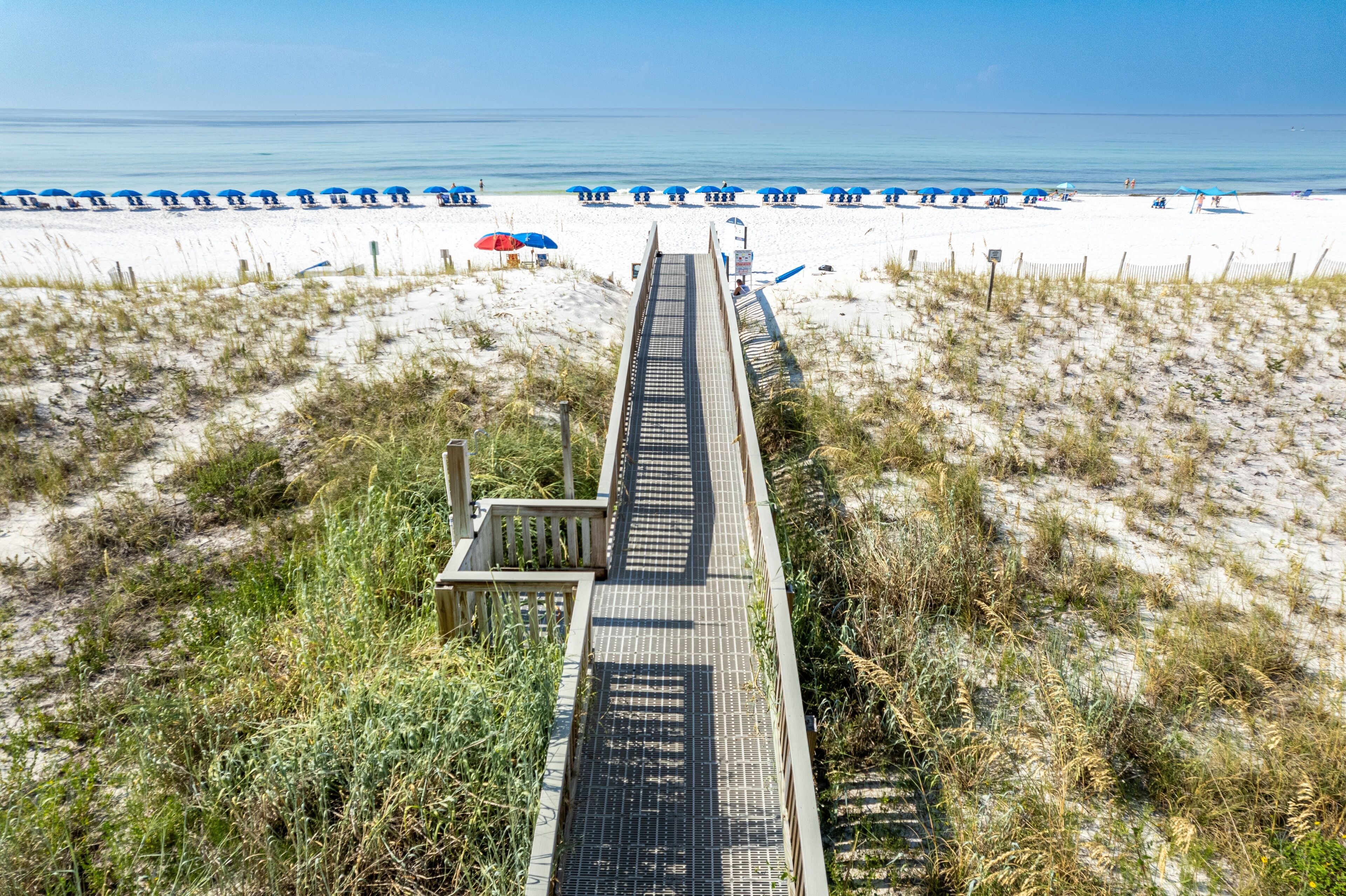 A wooden boardwalk leading to a beach with blue umbrellas and white sand.