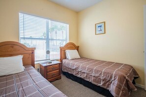 A bedroom with two single beds, a bedside table with a lamp, a window with blinds, and a framed picture on the wall. - 1415 Gulf Dr N Holmes Beach, Anna Maria Island 3 Bedroom Townhouse (Bradenton Beach)