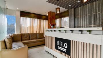 A hotel reception area with a reception desk, a sofa, and a large window with curtains.