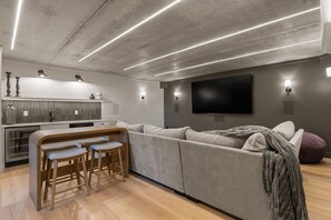 Een moderne woonkamer met een grijze bank, houten barstoelen, een flatscreen televisie en een keuken met een betegelde achterwand.