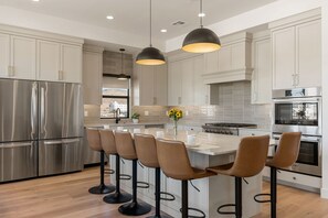 A modern kitchen with stainless steel appliances, a central island with bar stools, pendant lighting, and a view through the window. - Pickleball Penthouse Resort 21 Bedroom Home (St. George)