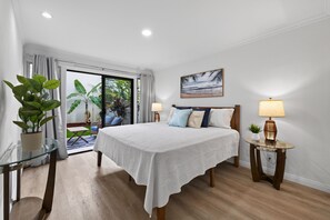 A hotel room with a bed, two bedside lamps, a small table with a lamp, a glass table with a plant, and a view of the outdoors through a sliding door. - Kona Coffee Villas F153 2 Bedroom Condo (Holualoa)