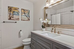 A bathroom with a toilet, sink, vanity, and two wall-mounted paintings. - Kona Coffee Villas F153 2 Bedroom Condo (Holualoa)