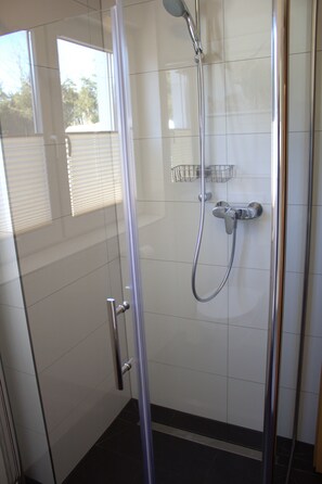 Une douche fermée par des parois en verre, dotée d’un pommeau de douche à main, d’un robinet mural et d’une petite étagère en métal.