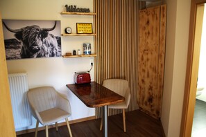 Une petite pièce avec une table en bois, une chaise, un radiateur, un tableau représentant un taureau, une horloge et une étagère sur laquelle est posé un panneau indiquant « Bienvenue ».
