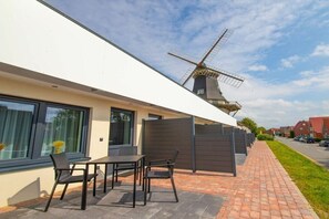 Ein modernes Gebäude mit einem Windrad im Hintergrund, einer Terrasse mit Stühlen und Tisch sowie einem Ziegelweg.