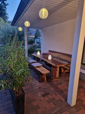 Een terras buiten met een houten tafel en banken, omringd door planten en een betegelde vloer.