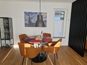 Een eethoek met een ronde tafel, vier stoelen, een hanglamp, een schilderij aan de muur en een raam met jaloezieën.