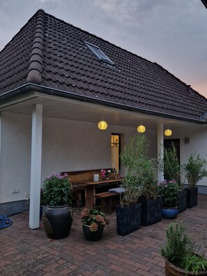 Ein Gebäude mit einem Ziegeldach, einer überdachten Terrasse mit einer Bank und Topfpflanzen sowie hängenden Laternen.