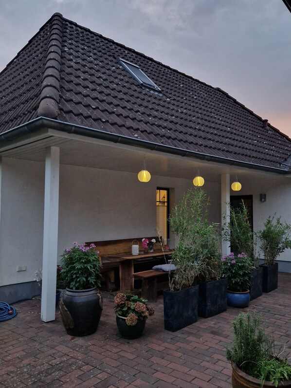 Een gebouw met een pannendak, een overdekte patio met een bankje en plantenbakken, en hangende lantaarns.
