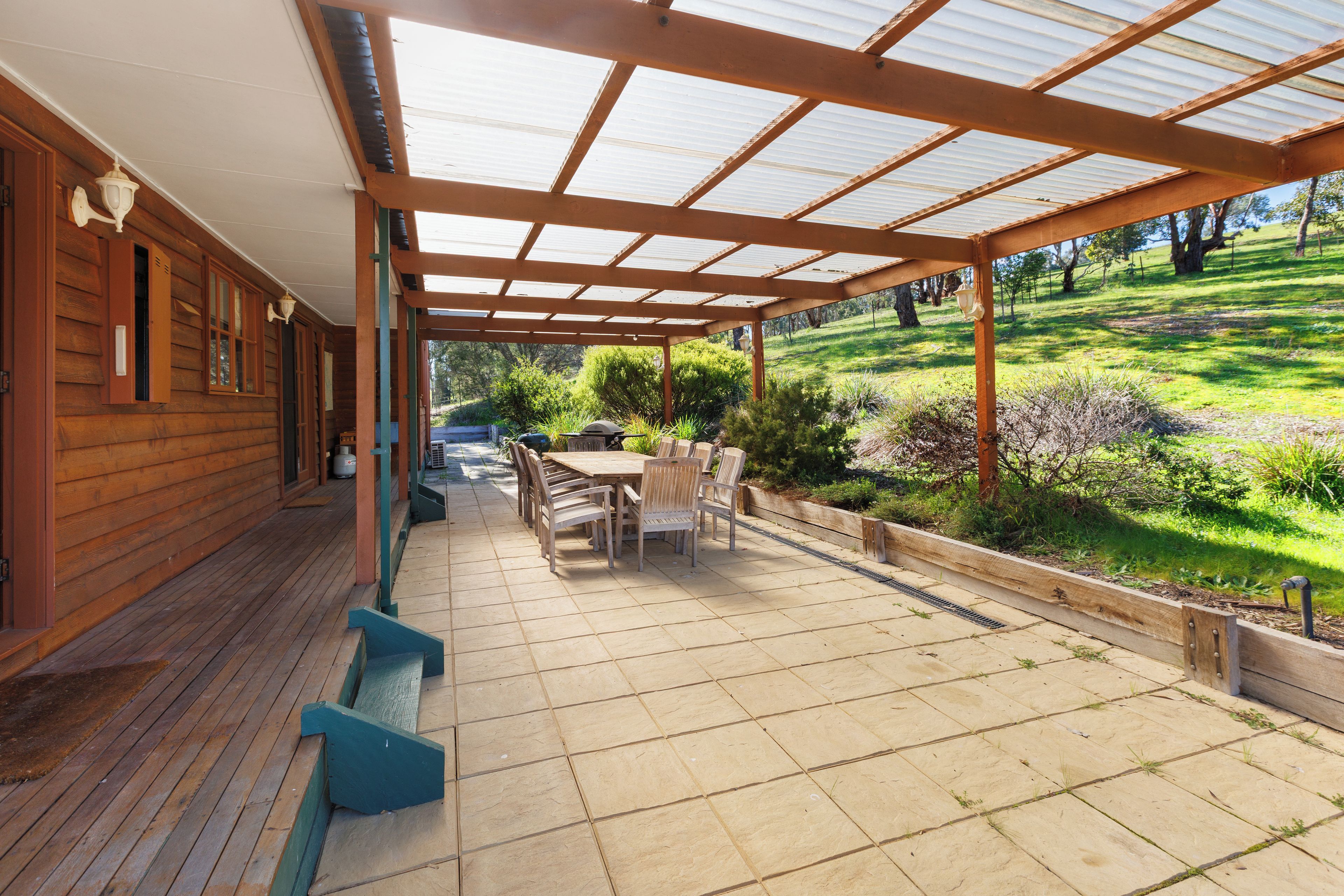 A covered patio with a wooden table and chairs, a grill, and a view of a grassy landscape.
