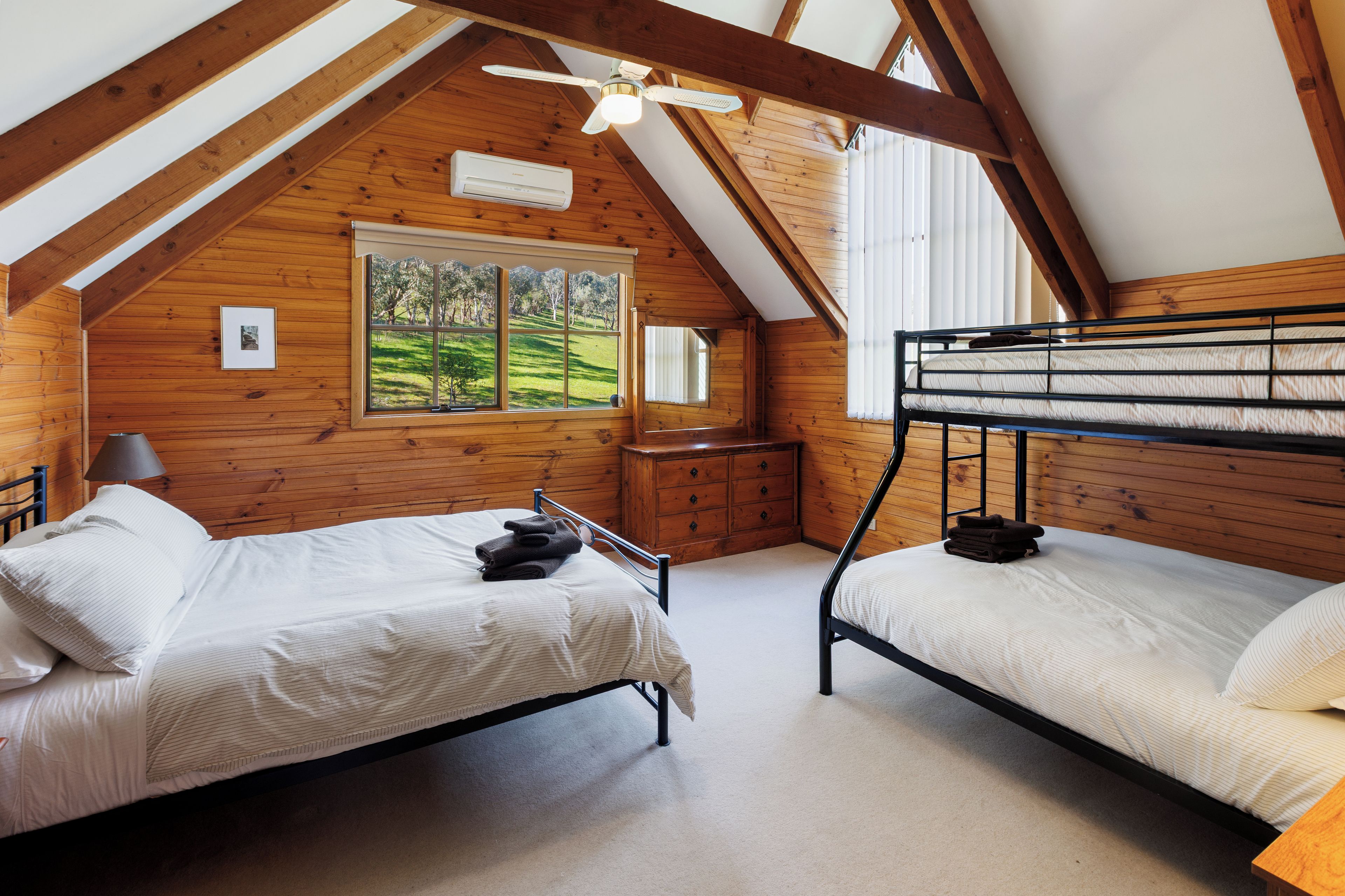 A bedroom with two bunk beds, a ceiling fan, a window with a view of a green landscape, and wooden walls.