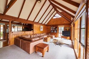 Een houten blokhut van binnen, met een houten plafond, een leren bank, een televisietoestel en een groot raam.