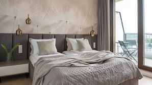 A modern bedroom with a large bed, a nightstand, a vase with plants, and a view of the city through a large window. - Bulvar Penthouse Collection Gdańsk (Gdańsk)