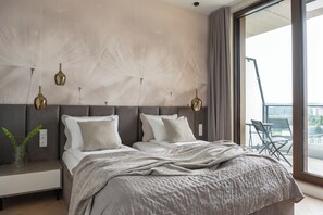 A modern bedroom with a large bed, a nightstand, a vase with plants, and a view of the city through a large window.