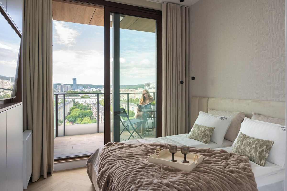 A bedroom with a large bed, a balcony with a city view, and a sliding glass door.