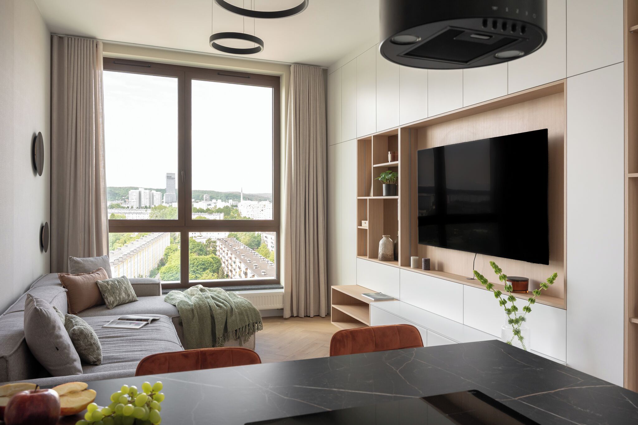 A modern living room with a large window, a flat-screen TV, a dining area, and a sofa.