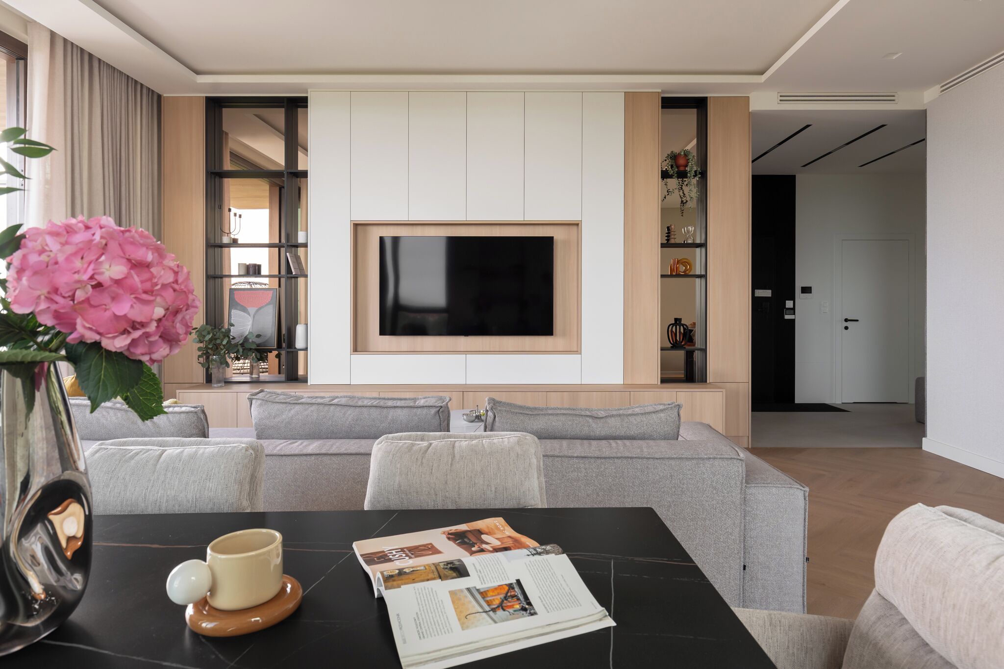 A modern living room with a flat-screen TV, grey sofas, a coffee table with a magazine, and a vase with pink flowers.