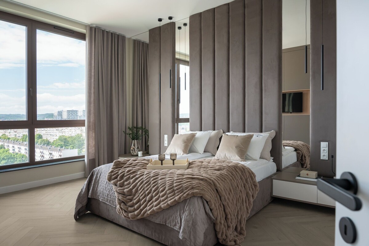 A modern bedroom with a large bed, a bedside table, a TV, and a view of the city.