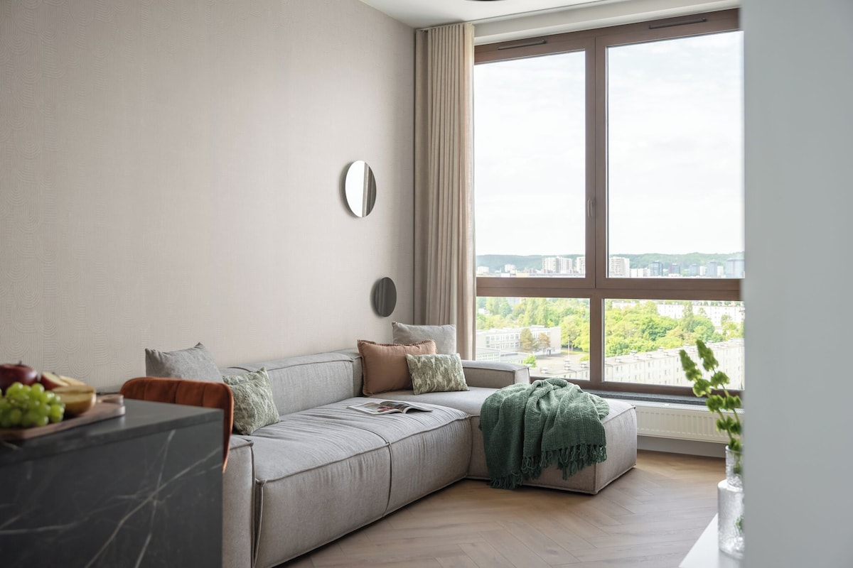 A modern living room with a grey sofa, a round mirror, and a view of a cityscape through large windows.