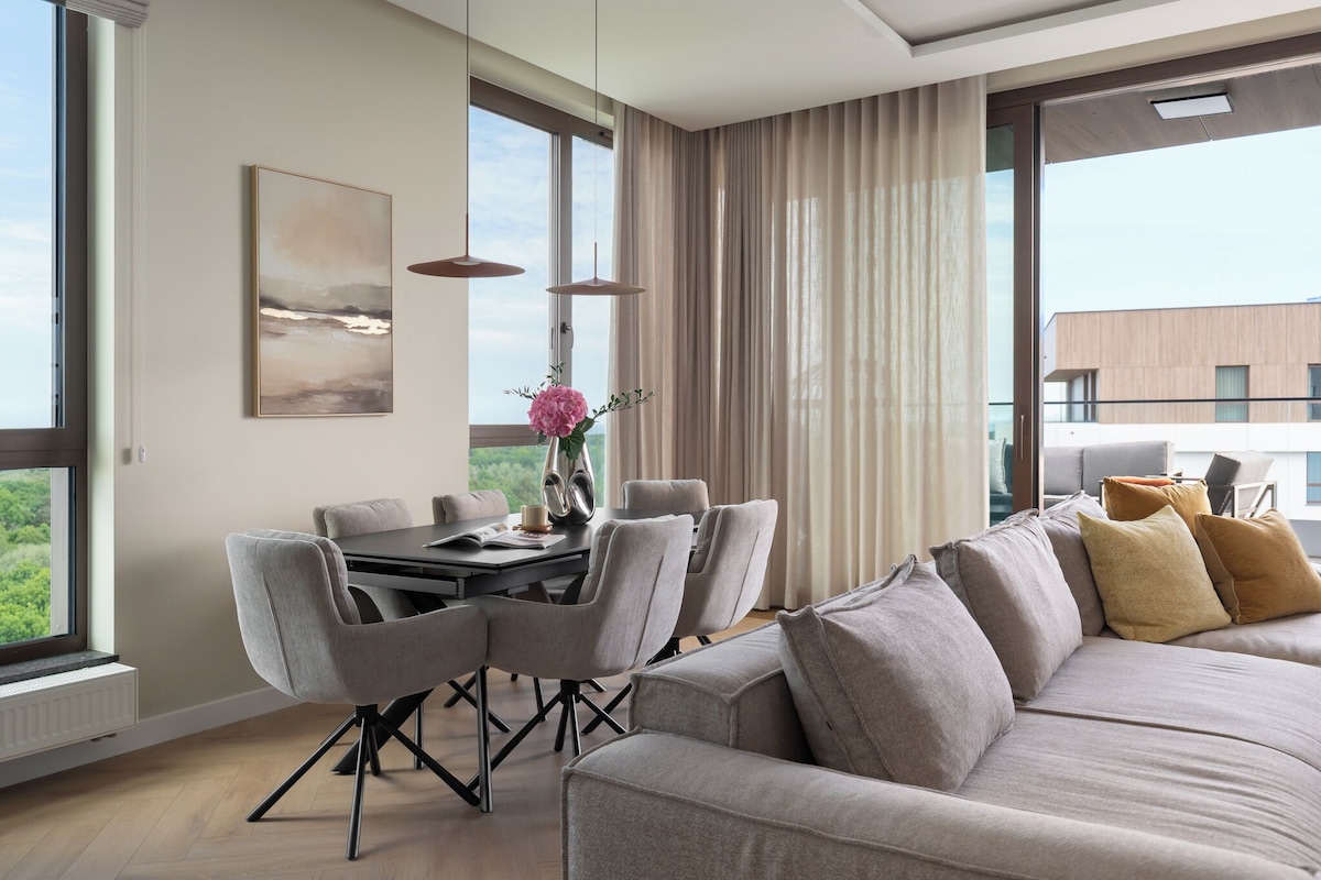 A modern living room with a sofa, dining table, chairs, and a view of greenery outside.
