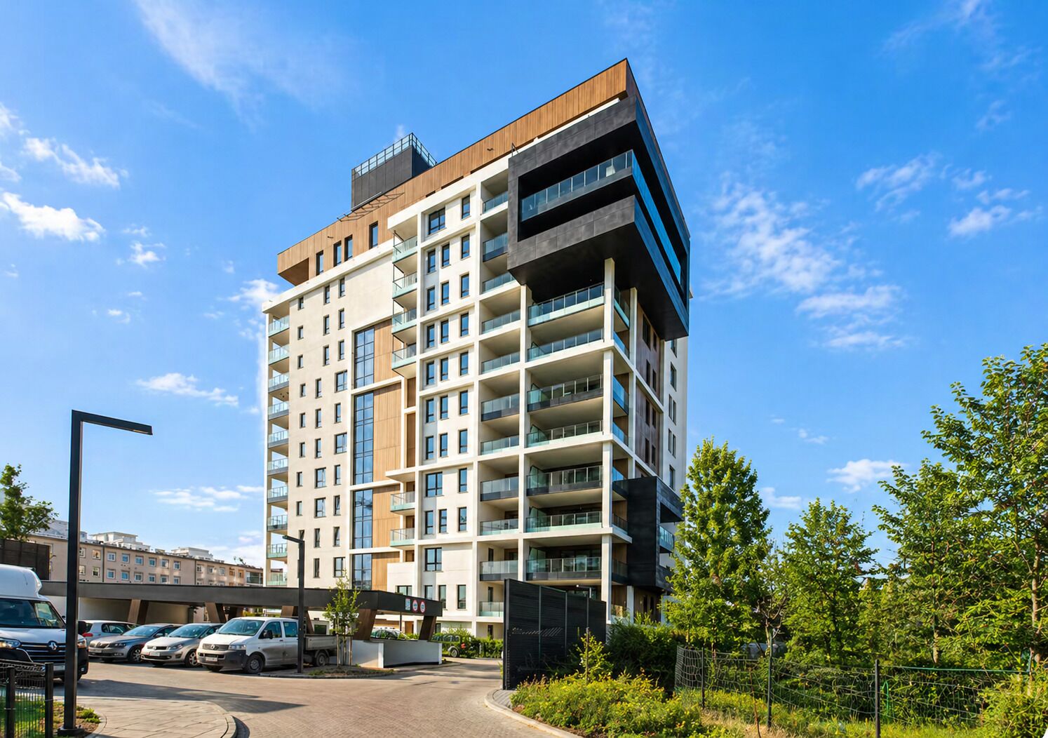 A multi-story residential building with balconies, a parking area, and a clear sky.