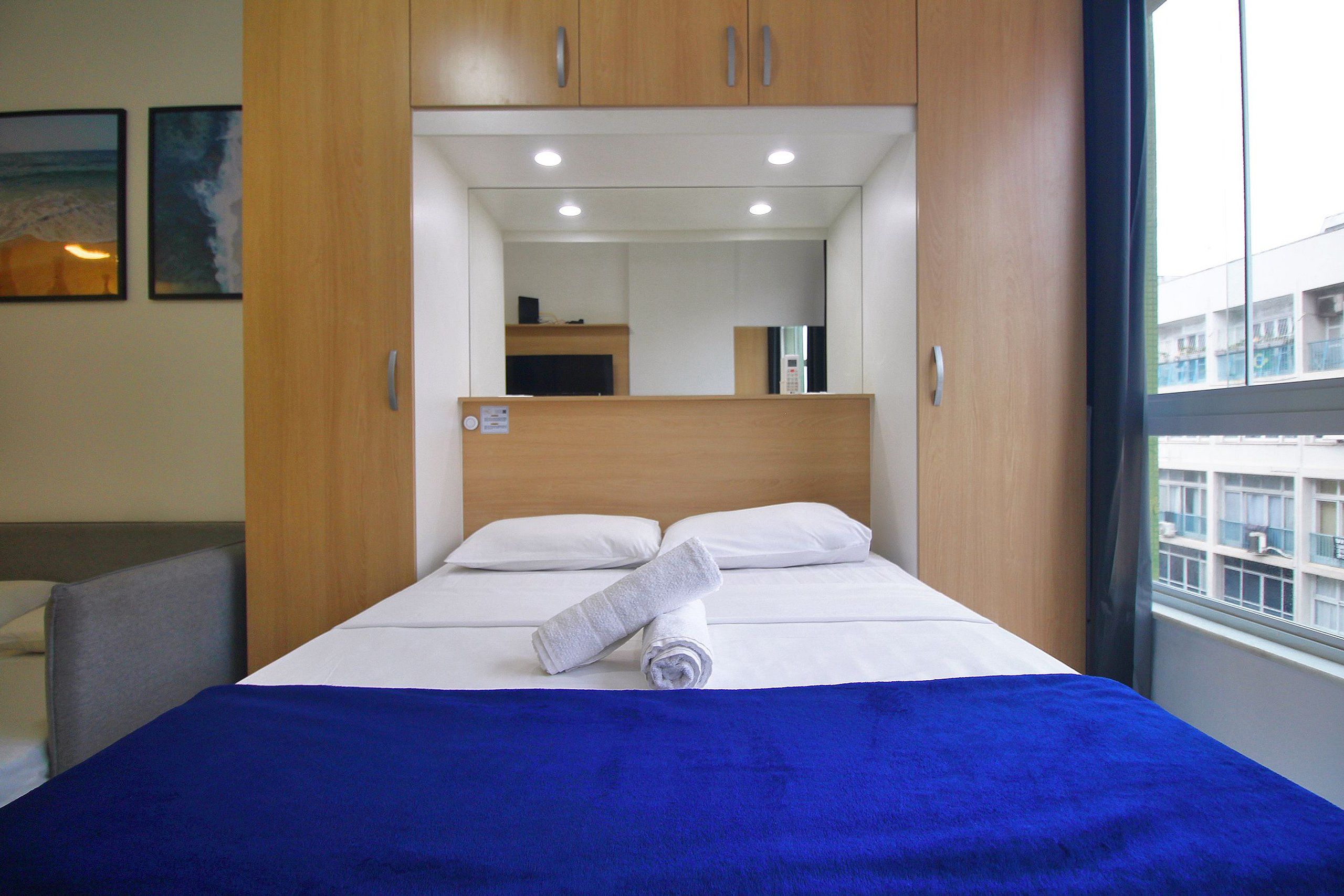 A hotel room with a bed, a blue blanket, a white pillow, a wooden headboard, and a window with a view of buildings.