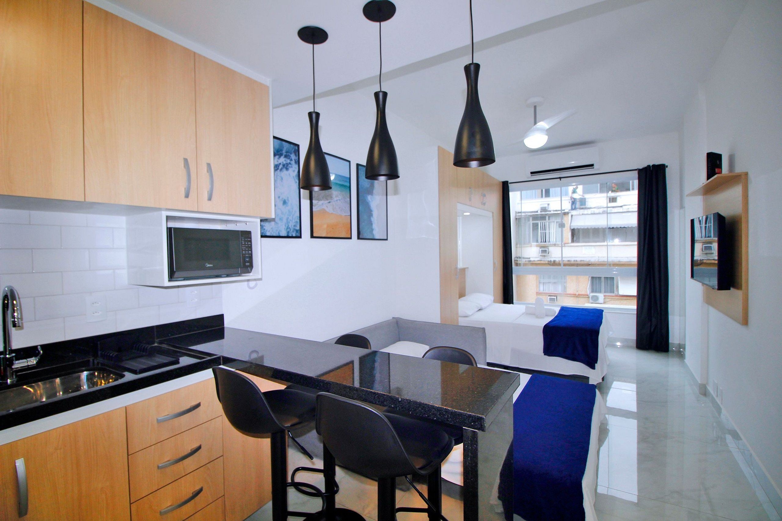 Een moderne keuken met een centraal eiland, zwarte barkrukken en een eettafel. Boven het eiland bevindt zich een magnetron, en de keuken heeft houten keukenkastjes.
