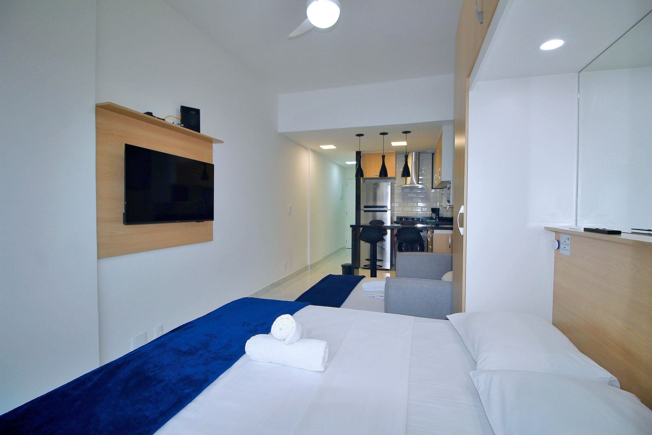A hotel room with a bed, a TV mounted on the wall, and a kitchenette area in the background.