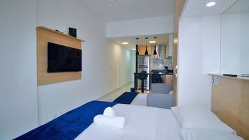 A hotel room with a bed, a TV mounted on the wall, and a kitchenette area in the background.