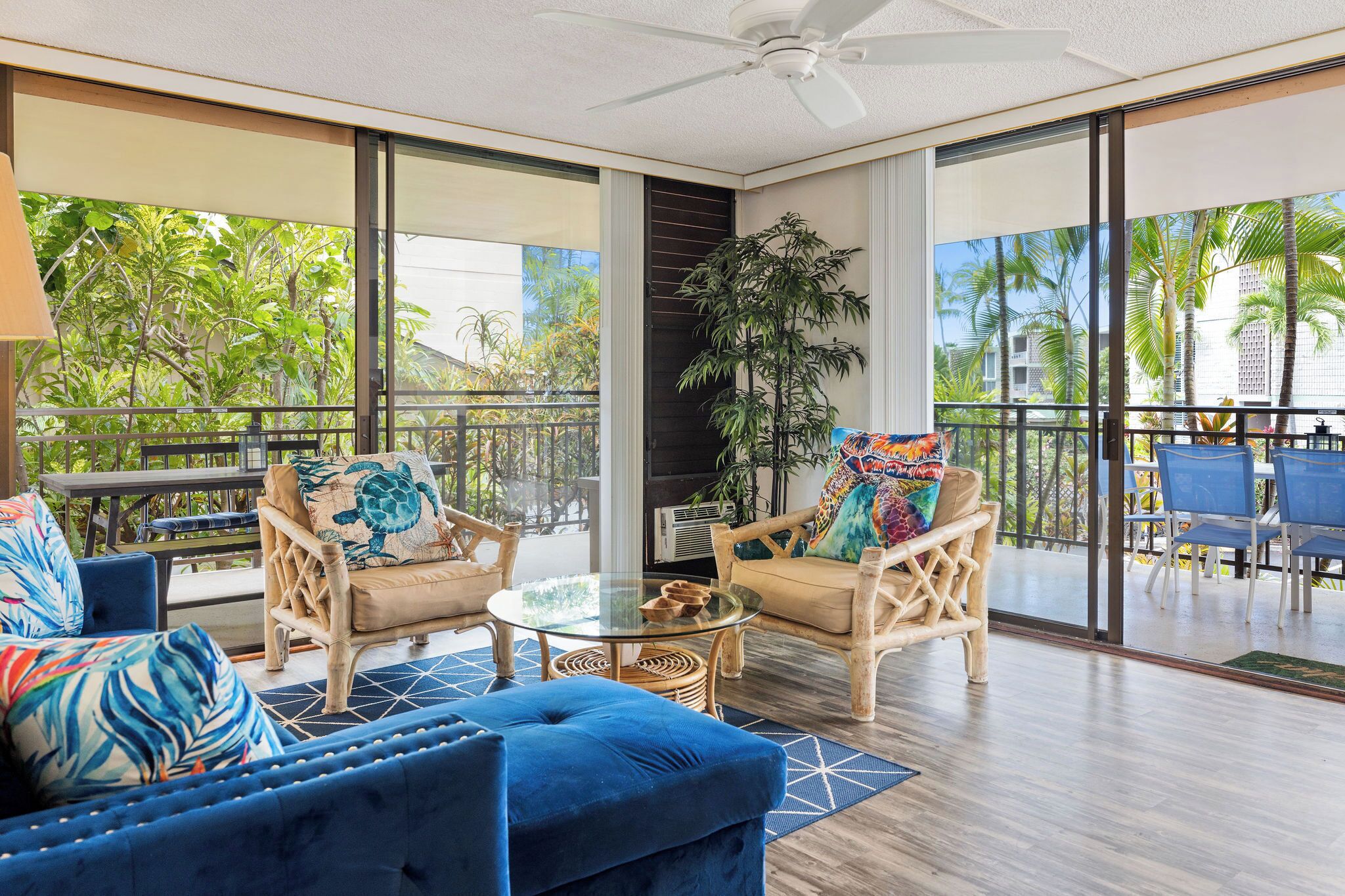 A spacious balcony with wicker furniture, a glass table, and a view of palm trees and buildings.
