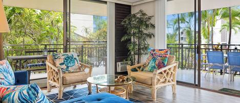 A spacious balcony with wicker furniture, a glass table, and a view of palm trees and buildings.