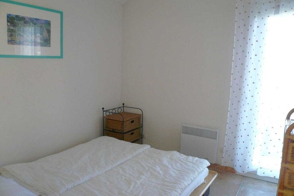 Een eenvoudige kamer met een bed, een radiator, een raam met gordijnen en een lijst met een afbeelding aan de muur.