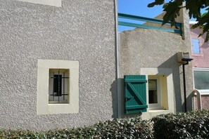 A building with a textured facade, a window with a metal grille, and a smaller window with green shutters. - Le Clos d'Elsa N° 19 (Gruissan)