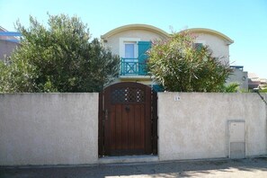 A gated entrance with a wooden gate, a white wall, and a building with a balcony in the background. - Le Clos d'Elsa N° 19 (Gruissan)