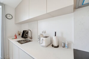 A modern kitchen with white cabinetry, a sink, a toaster, a kettle, and a clock on the wall. - Balcony Studio in Northfields - Apt 1 (London)