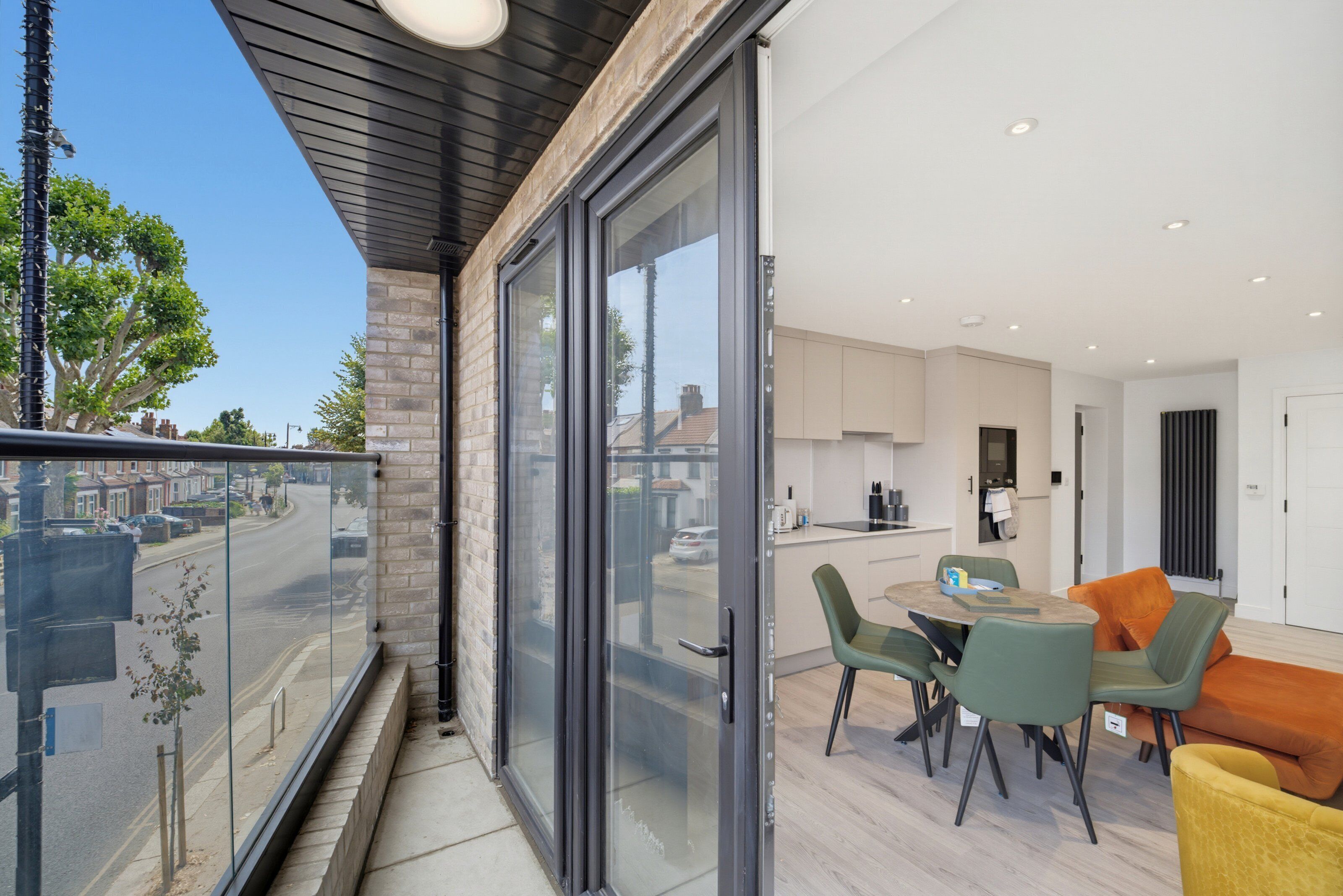 A modern apartment with a balcony overlooking a street, a kitchen with contemporary appliances, and a dining area with green and orange chairs.
