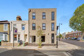 A modern two-story building with a brick facade, a small front garden, and a parking area with cars. - Balcony Studio in Northfields - Apt 1 (London)
